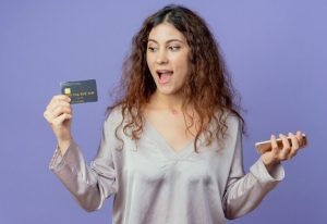 New Home 34 Mulher jovem segurando um celular em uma mão e um cartão de crédito na outra, demonstrando entusiasmo ao receber a aprovação de um cartão de crédito sem anuidade, representando liberdade financeira e praticidade no dia a dia.