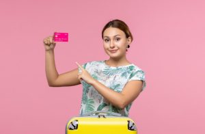 Homepage 7 Mulher sorridente segurando um cartão rosa e apontando para ele, ao lado de uma mala amarela, representando os benefícios dos cartões de crédito com bônus de milhas para quem gosta de viajar.