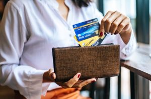 Homepage 5 Mulher retira cartões de crédito sem anuidade de uma carteira marrom enquanto está sentada próxima a uma janela, simbolizando praticidade, organização financeira e liberdade no uso de serviços bancários isentos de taxas.