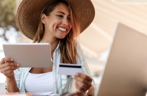 Homepage 6 Mulher sorridente usando chapéu, segurando um cartão de crédito e um tablet enquanto realiza transações online, simbolizando o uso inteligente de programas de milhas para compras e viagens.
