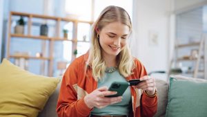 Mulher usando celular e cartão em casa — ilustração de como escolher um banco digital com segurança e conveniência.