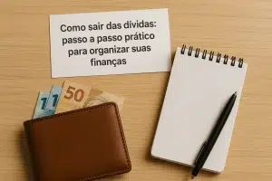Folha de papel com o título 'Como sair das dívidas: passo a passo prático para organizar suas finanças', posicionada sobre uma mesa de madeira clara. Ao lado, há uma carteira marrom aberta com cédulas de reais parcialmente visíveis (uma de R$50 e duas de R$2), um bloco de notas quadriculado e uma caneta preta.