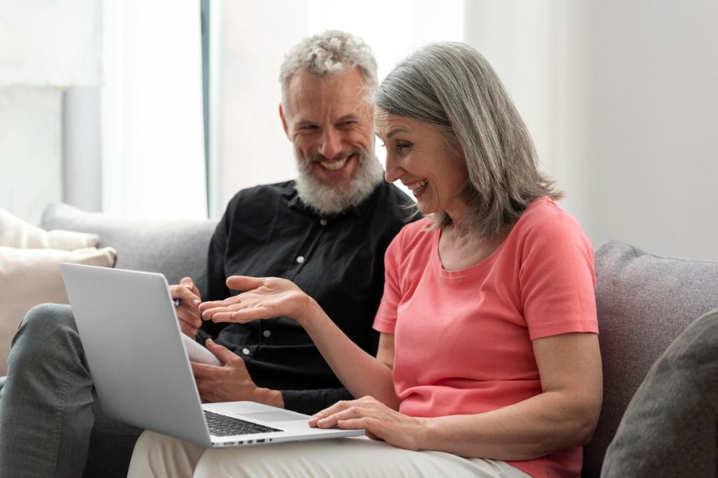 Casal de idosos sorridente sentado no sofá usando um notebook, demonstrando satisfação ao pesquisar opções de empréstimo para negativado consignado INSS em casa.