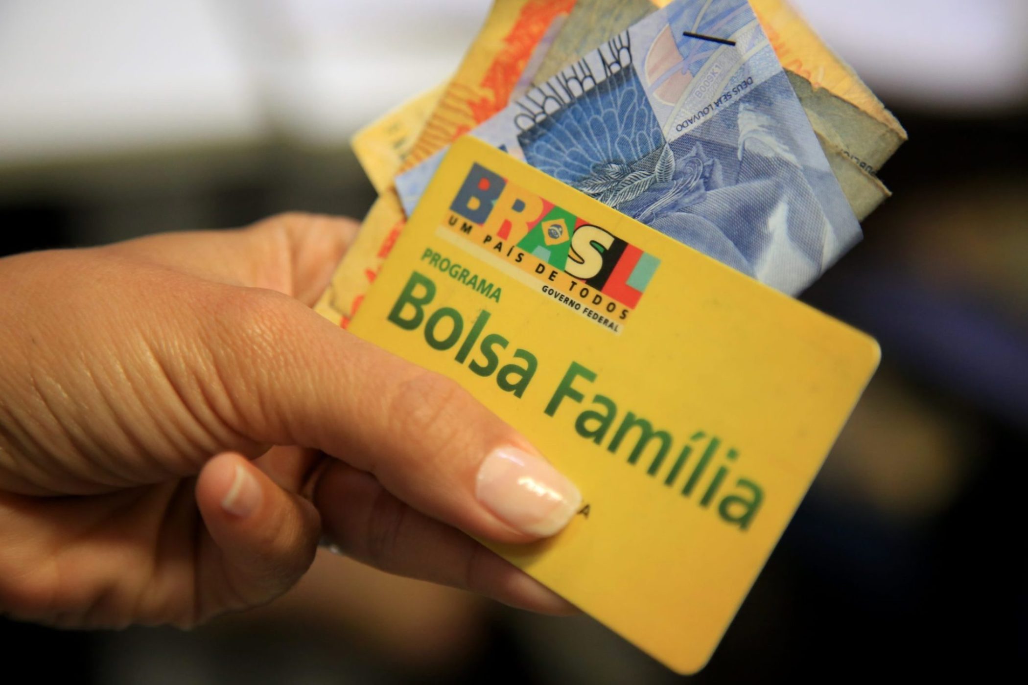 Pessoa segurando o cartão do programa Bolsa Família junto a cédulas de real, simbolizando o recebimento do benefício social.
