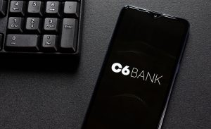 Smartphone exibindo a tela inicial do aplicativo C6 Bank sobre uma mesa preta, ao lado de um teclado de computador.