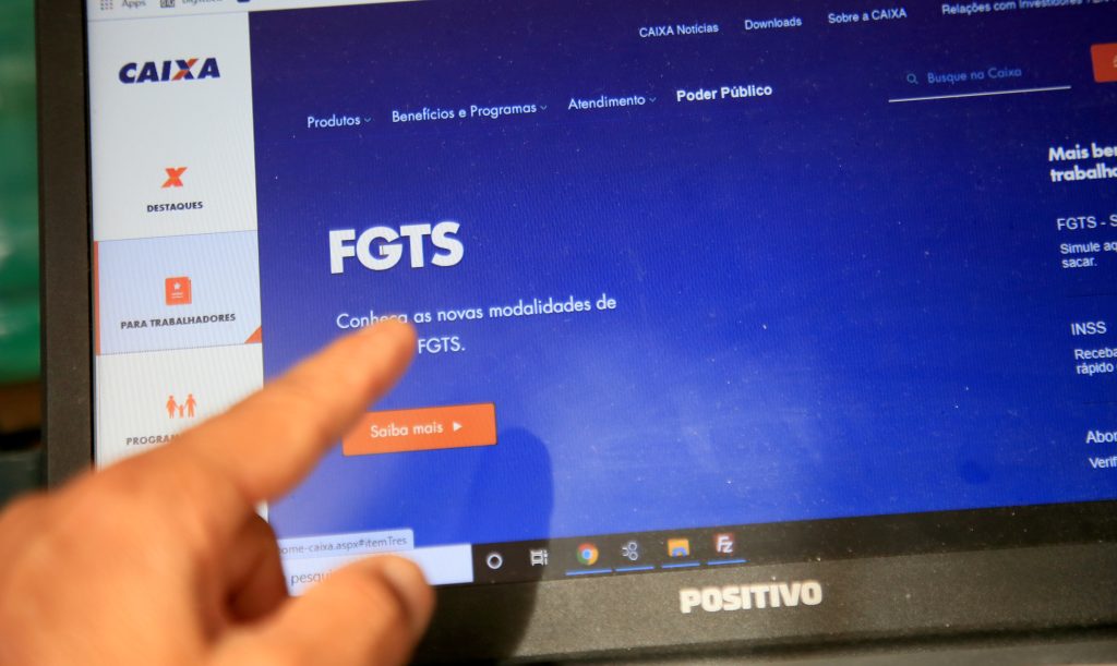 Tela de notebook exibindo o site oficial da Caixa Econômica Federal, com destaque para a seção do FGTS (Fundo de Garantia do Tempo de Serviço). Um dedo aponta para a frase “Conheça as novas modalidades de FGTS”. A interface mostra botões de navegação lateral e o botão laranja “Saiba mais”.