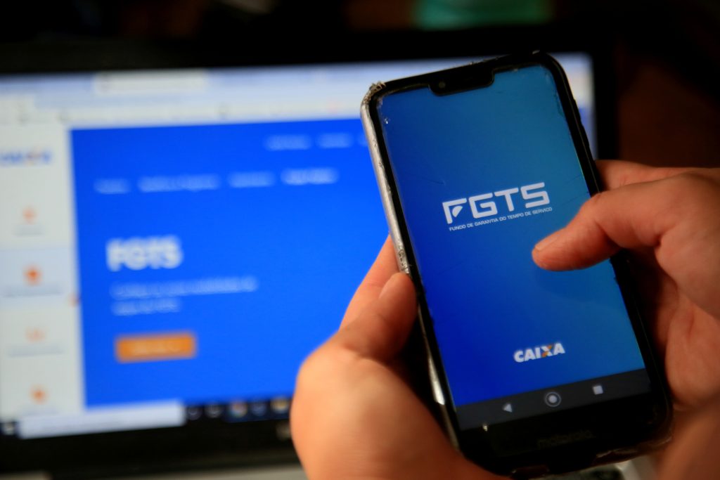 Saque FGTS: modalidades, como solicitar e quando vale a pena usar. 1 Pessoa segurando um celular com a tela aberta no aplicativo do FGTS (Fundo de Garantia do Tempo de Serviço), da Caixa Econômica Federal. Ao fundo, no computador, aparece a tela inicial do site do FGTS. A foto representa consulta ou movimentação de valores do fundo pelo app oficial para saque de FGTS.