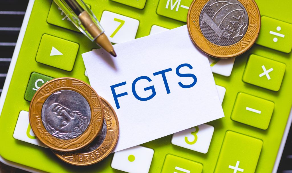 Calculadora verde com algumas moedas de um real ao redor e um papel branco ao centro com a sigla “FGTS” em destaque azul. Na parte superior, uma caneta dourada também aparece parcialmente. A imagem transmite a ideia de cálculos, finanças e controle do Fundo de Garantia do Tempo de Serviço.