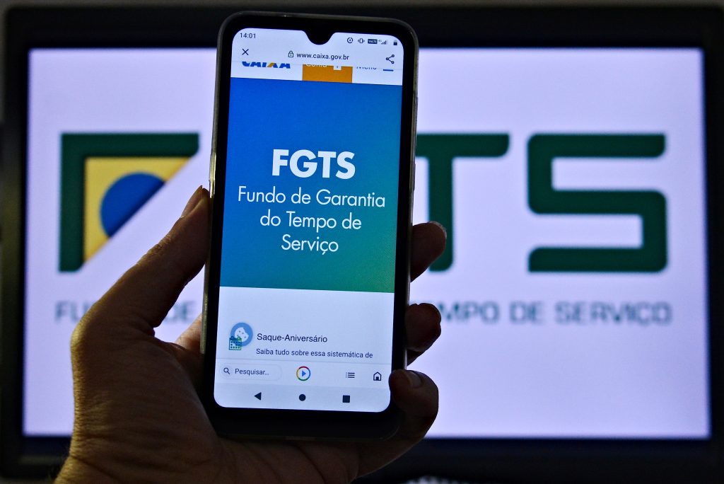 Pessoa segurando um celular com a tela aberta no aplicativo do FGTS (Fundo de Garantia do Tempo de Serviço), da Caixa Econômica Federal. Ao fundo, no computador, aparece a tela inicial do site do FGTS. A foto representa consulta ou movimentação de valores do fundo pelo app oficial.
