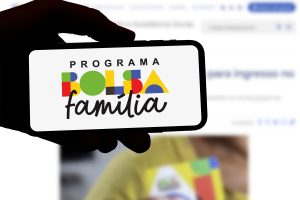Pessoa segurando um celular que exibe o logotipo atualizado do Programa Bolsa Família, com letras coloridas e formas geométricas, em destaque sobre fundo branco. Ao fundo, desfocado, está uma tela de computador com informações relacionadas ao programa.