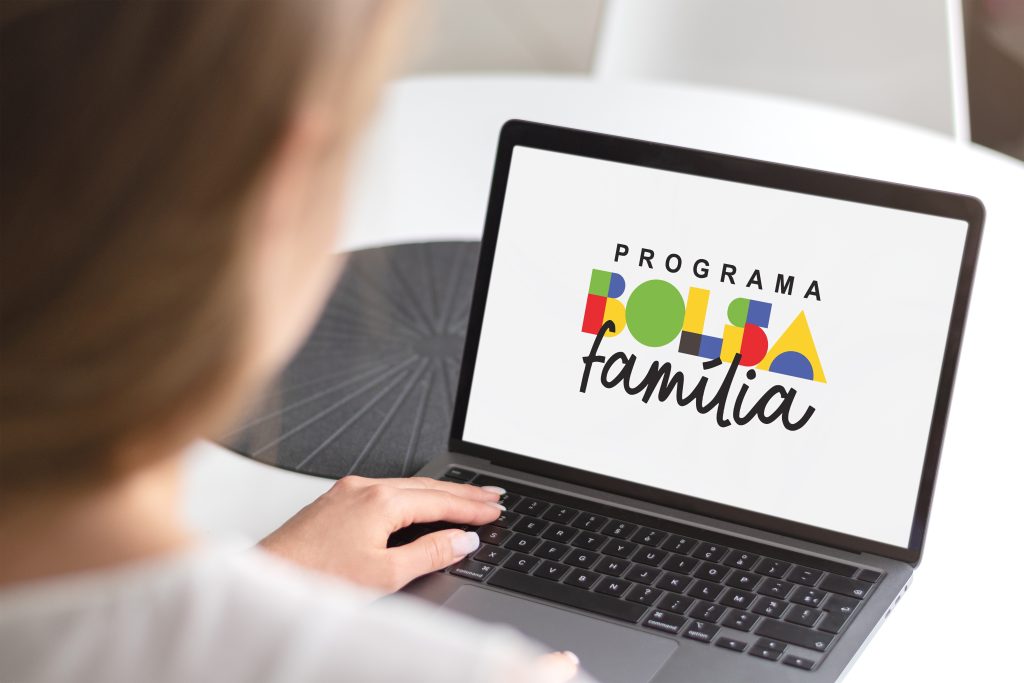 Mulher utilizando um notebook em uma mesa. Na tela aparece o logotipo do Programa Bolsa Família em letras coloridas e fundo branco. A cena transmite a ideia de consulta ou cadastro no benefício por meio digital.