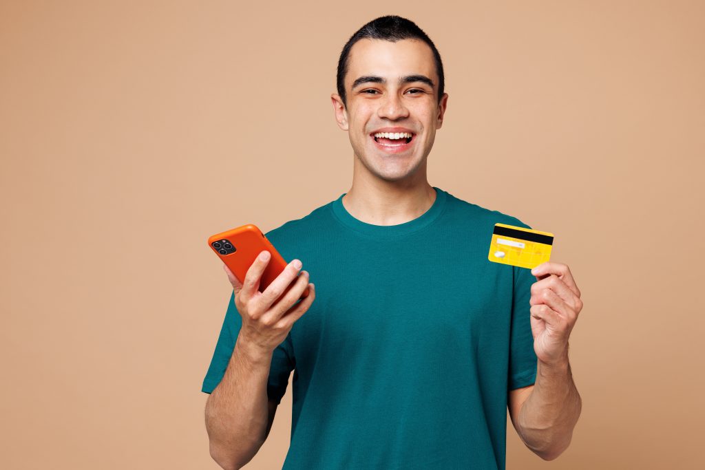 Cartão de crédito com cashback sem anuidade: melhores opções em 2025 2 Homem sorrindo enquanto segura o celular e um cartão de crédito amarelo, simbolizando praticidade ao usar um cartão de crédito com cashback sem anuidade.