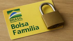 Cartão amarelo do programa Bolsa Família do Governo Federal sobre uma mesa de madeira, com um cadeado metálico apoiado por cima para representar a situação de Bolsa Família bloqueado e a suspensão temporária do benefício.
