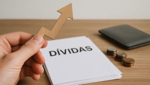 Mão segurando uma seta de madeira apontando para cima em frente a um maço de papéis com a palavra “Dívidas”, representando o aumento do endividamento e a busca por soluções de como sair das dívidas em 2025.