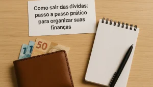 Folha de papel com o título 'Como sair das dívidas: passo a passo prático para organizar suas finanças', posicionada sobre uma mesa de madeira clara. Ao lado, há uma carteira marrom aberta com cédulas de reais parcialmente visíveis (uma de R$50 e duas de R$2), um bloco de notas quadriculado e uma caneta preta.