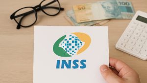 Documento com o logotipo do INSS sendo segurado por uma mão sobre mesa de madeira clara, com óculos, notas de reais e calculadora ao fundo, representando ambiente de análise financeira e explicação sobre empréstimo consignado INSS como funciona.