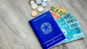 Carteira de Trabalho azul ao lado de notas de real e moedas sobre superfície de madeira, representando pagamento do abono salarial PIS/PASEP.