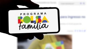 Pessoa segurando um celular que exibe o logotipo atualizado do Programa Bolsa Família, com letras coloridas e formas geométricas, em destaque sobre fundo branco. Ao fundo, desfocado, está uma tela de computador com informações relacionadas ao programa.